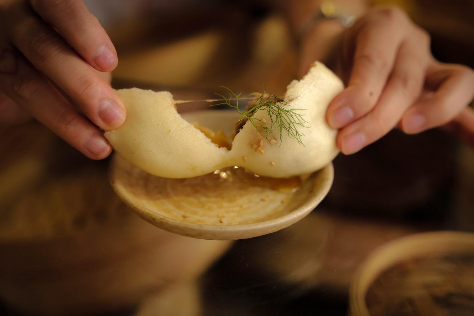 Two hands pull a grilled bao open over a small ceramic dish, with dill and sesame.