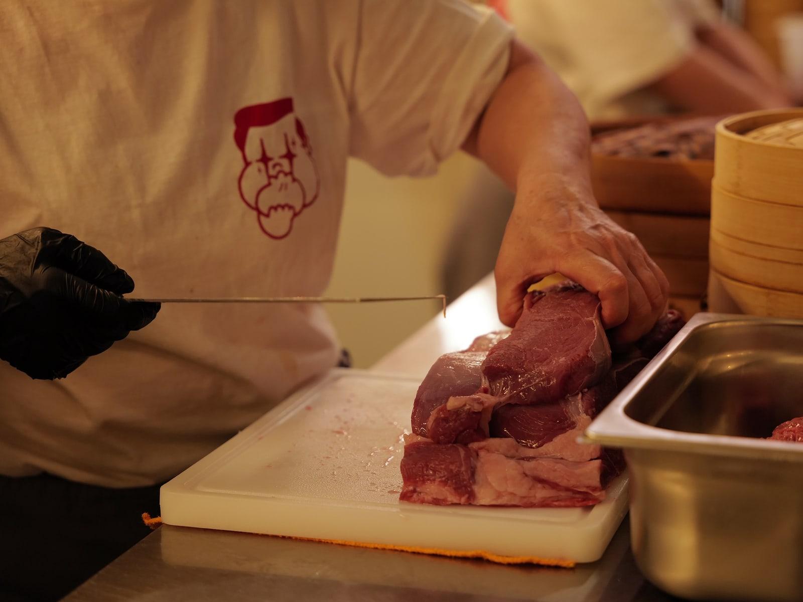 Beef being trimmed in the kitchen, the Bao Bros mascot printed on the cook's T-shirt, bamboo steamers beside it.